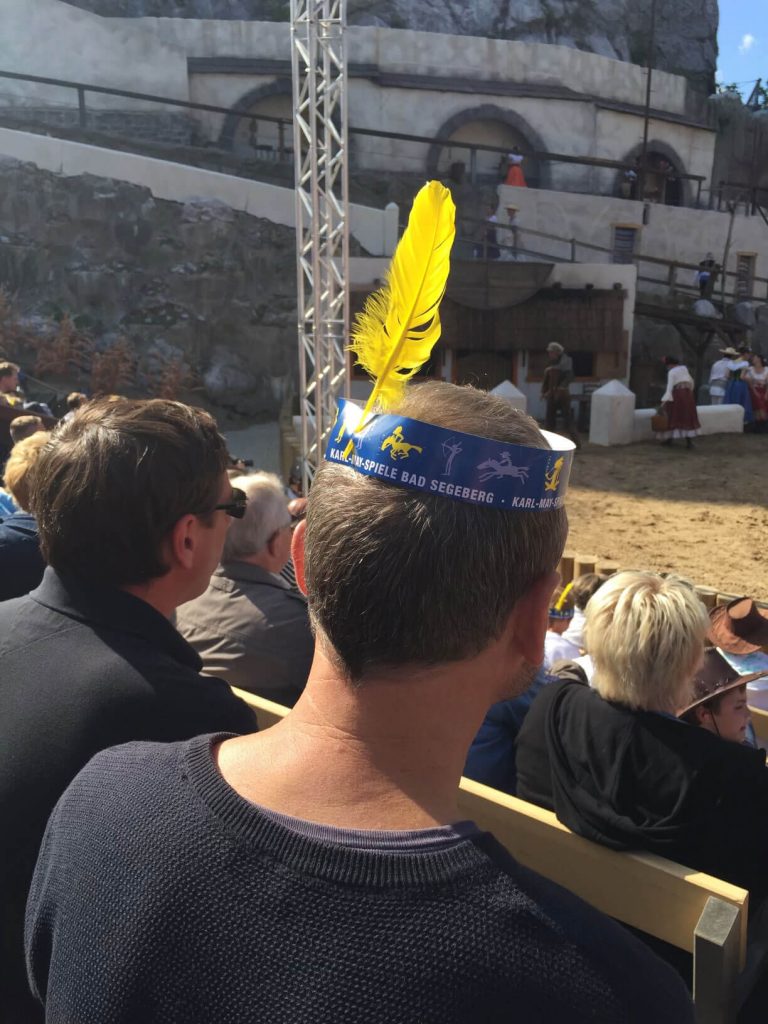 A photo of the back of a man's head, wearing a novelty paper crown with a bright yellow feather stuck out the top.