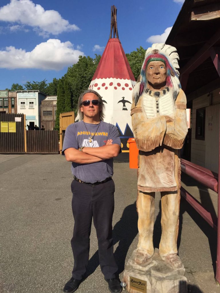 Drew stands with his arms crossed next to a wood carved statue depicting an Indigenous person in full headdress.