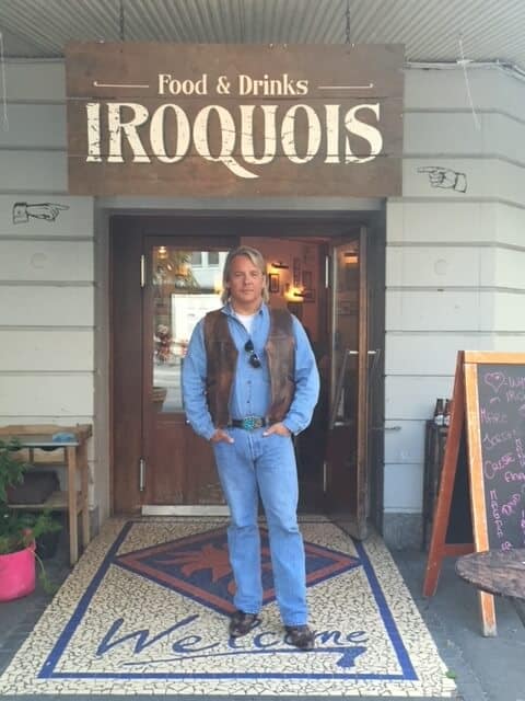 Drew stands in front of a sign that says "food and drinks IROQUIS"