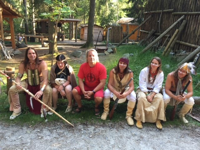 Drew is sitting on a long bench surrounded by 5 other people all dressed in Indigenous outfits