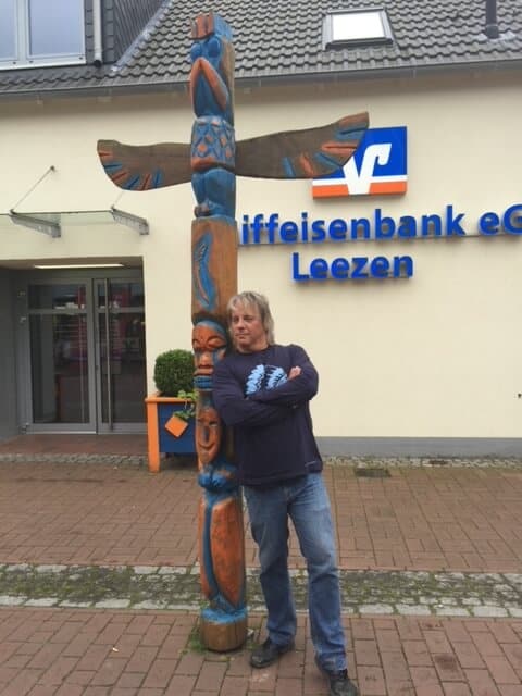 Drew stands in front of a German totem pole, arms crossed and leaning onto it