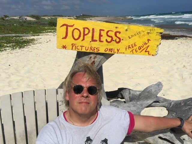 Drew sits on a beach in fornt of a sign that reads "TOPLESS PICTURES ONLY- FREE TEQUILA!"
