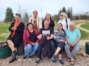 Drew sits outside in a group with 7 other Indigenous writers, men and women of different ages, all smiling for the camera