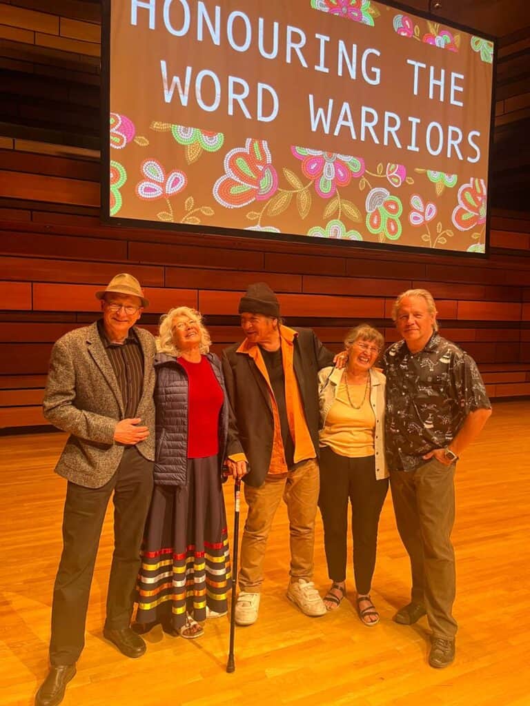 Drew Taylor stands in a line of authors, all laughing and smiling like old friends, on stage with a presentation screen behind that reads "Honouring the Word Warriors"