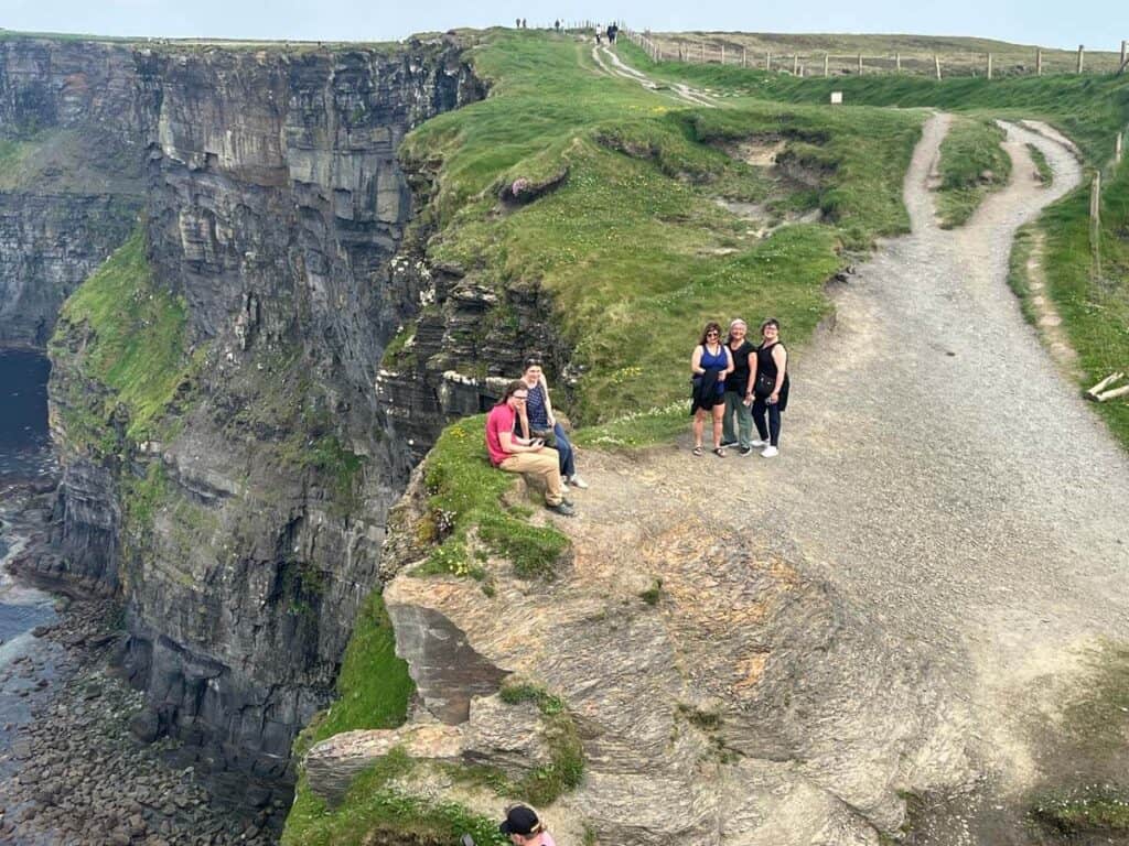 A far away shot of a rugged landscape, with a sheer cliff face on the left and a winding path to the lookout on the right. A group of hikers sits near the cliff edge and smile for the camera
