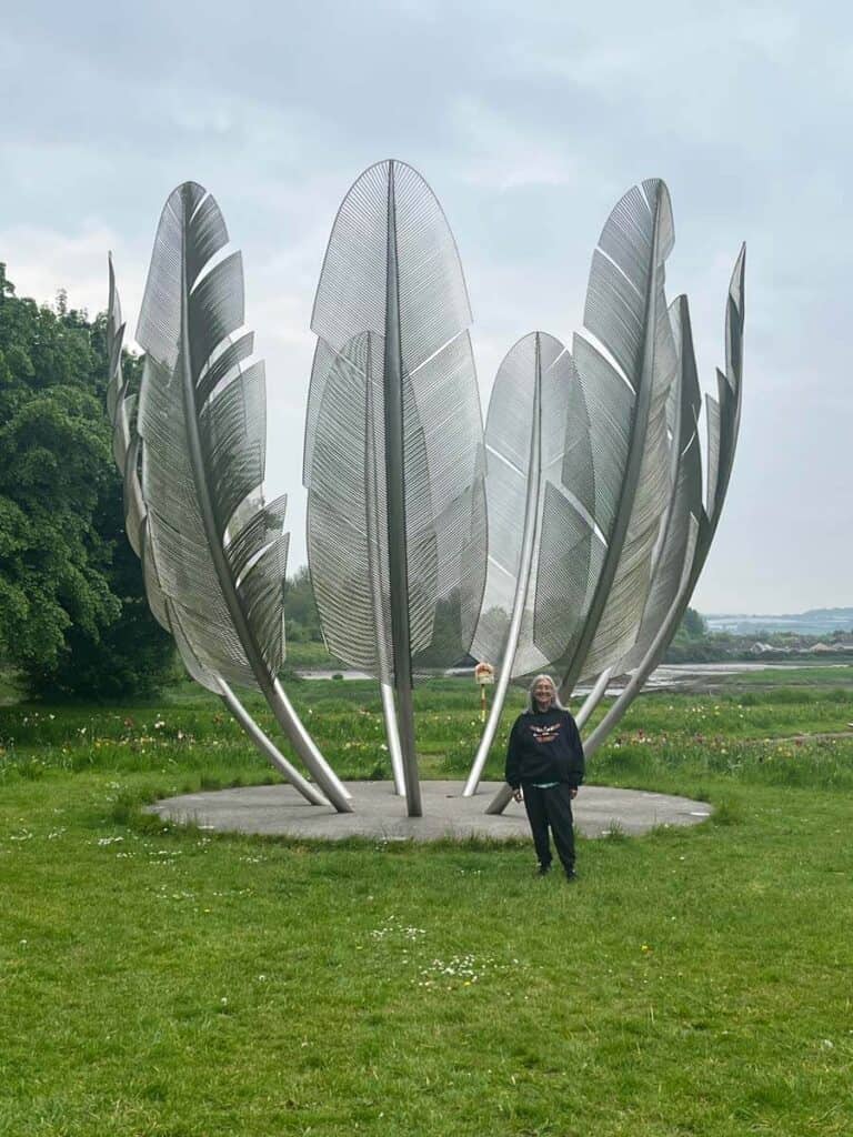 A woman stands in front of an outdoor sculpture, a series of giant feathers standing in a circle with their shafts stuck into the ground, all made of metal.