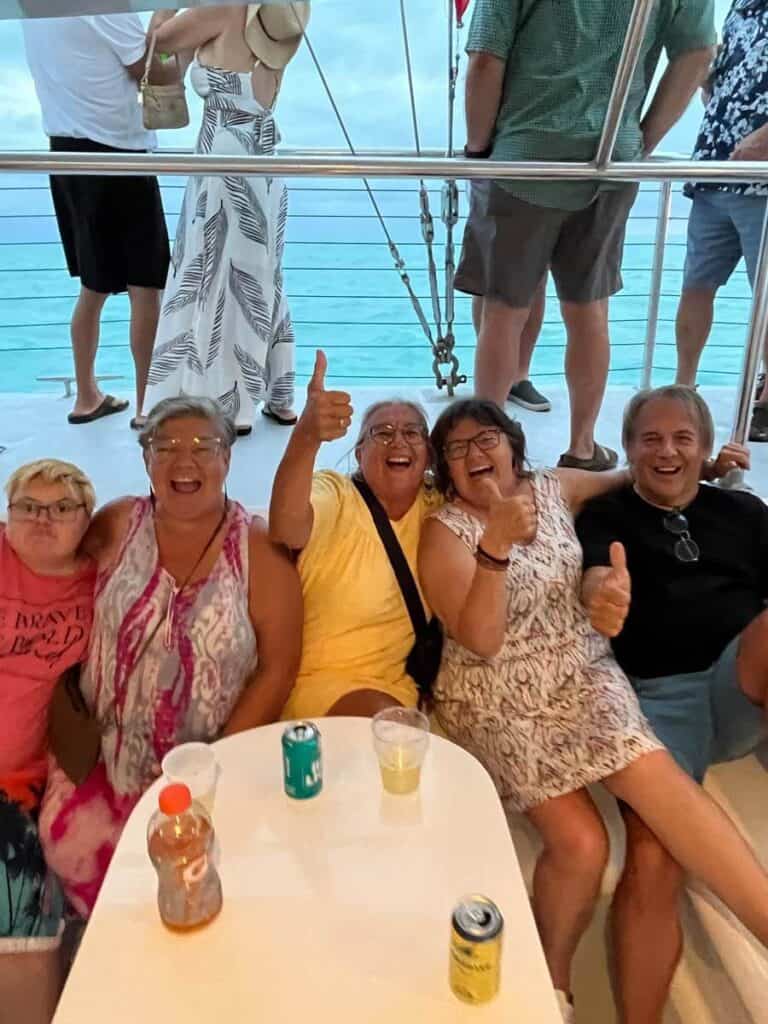 A happy group of friends are sitting shoulder to shoulder around a small table with drinks, they are all smiles and giggles, giving thumbs up for the camera.