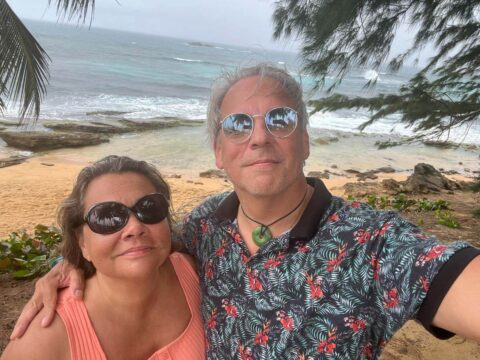 Drew and his partner, Janine, take a vacation selfie, both in summer shirts with a tropical beach behind them