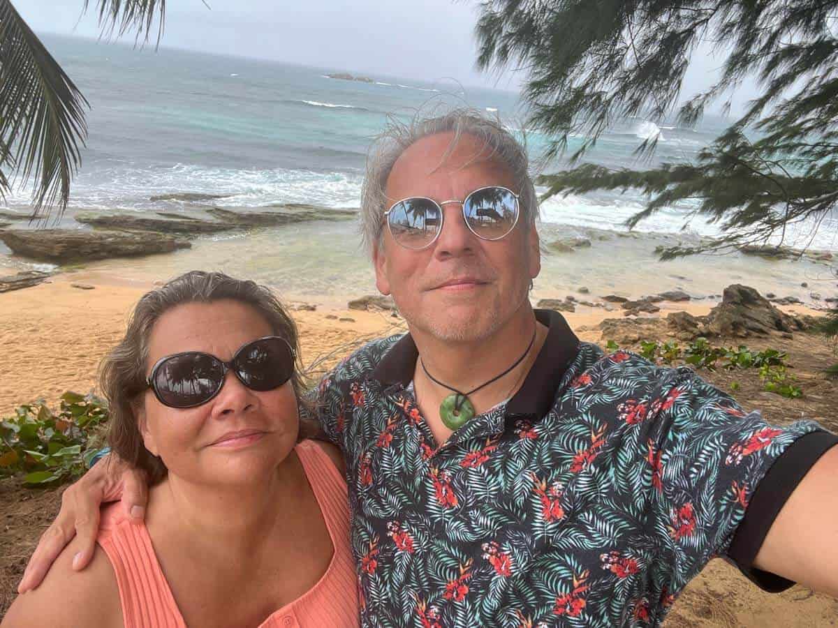 Drew and his partner, Janine, take a vacation selfie, both in summer shirts with a tropical beach behind them